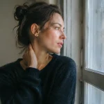 Femme porte la main au cou devant une fenetre
